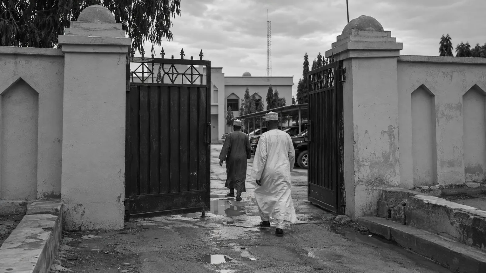 Two figures enter an administrative compound in Kano, Nigeria, beneath an overcast sky. (AI-generated image / EconoTimes)