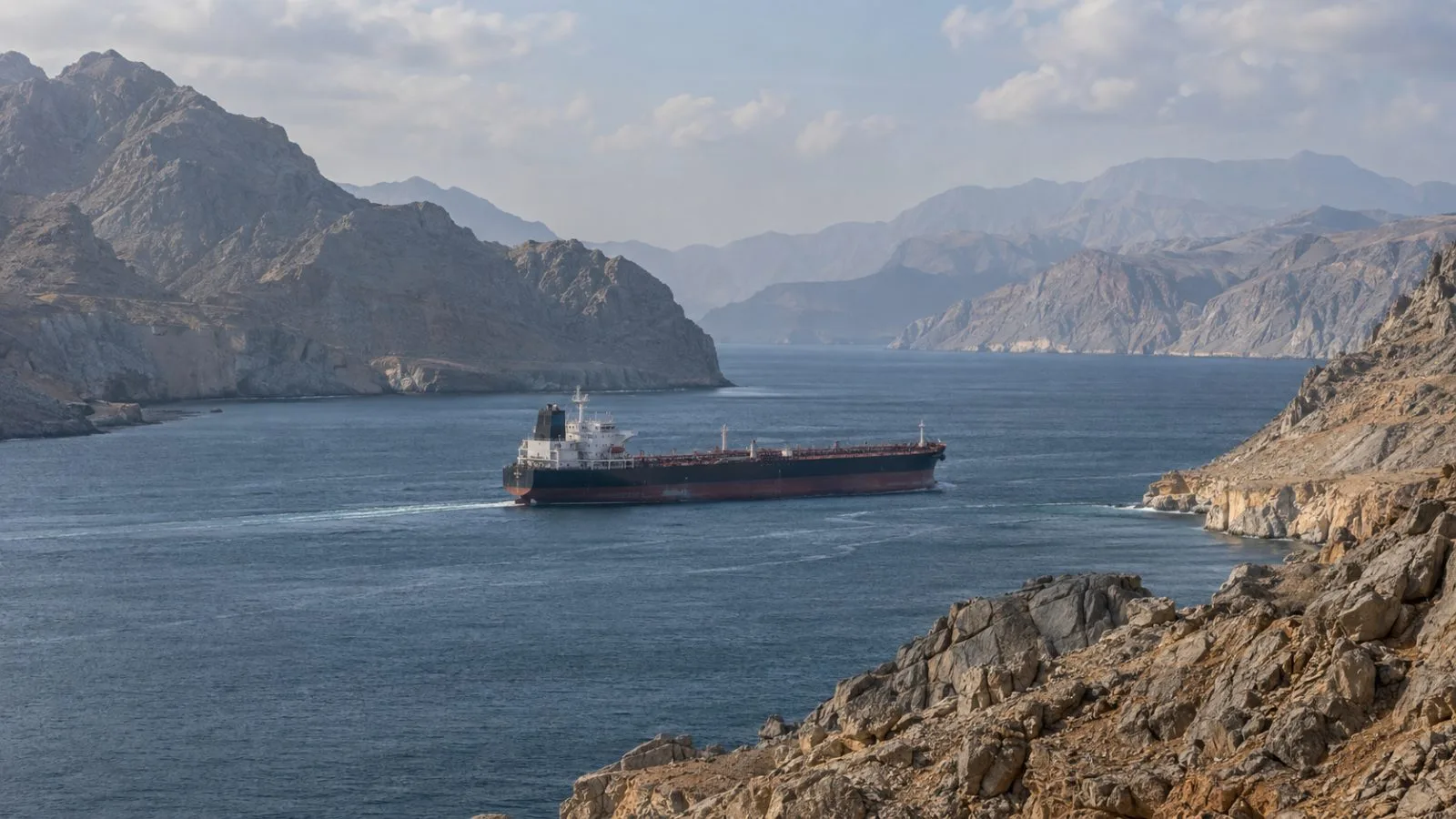 A tanker transits the Strait of Hormuz near Khasab, Oman, in hazy morning light (AI-generated image / EconoTimes)