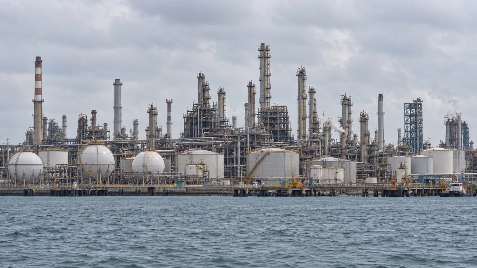 Refinery pipes and storage tanks stand on Pulau Bukom, Singapore, beneath an overcast sky. / EconoTimes.ai