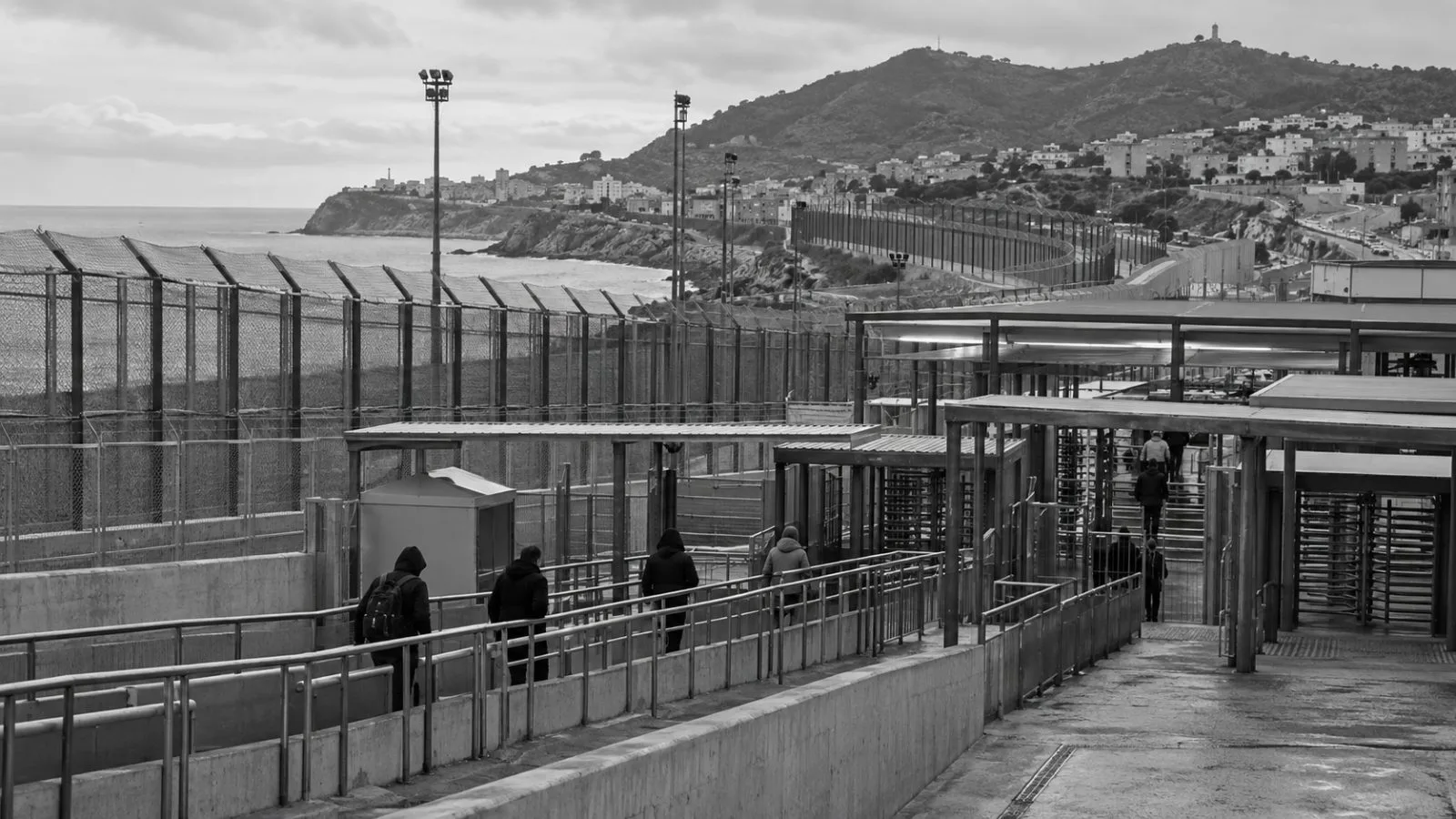 Migrants wait near a border fence at Ceuta’s crossing with Morocco, Spain / EconoTimes.ai (mono)